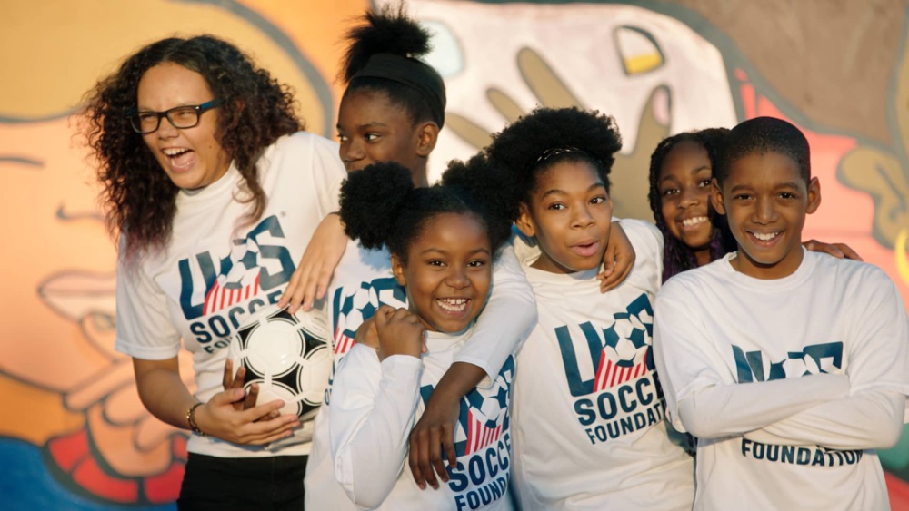 Kids smiling with their arms around each other in soccer jerseys and one girl is holding a soccer ball